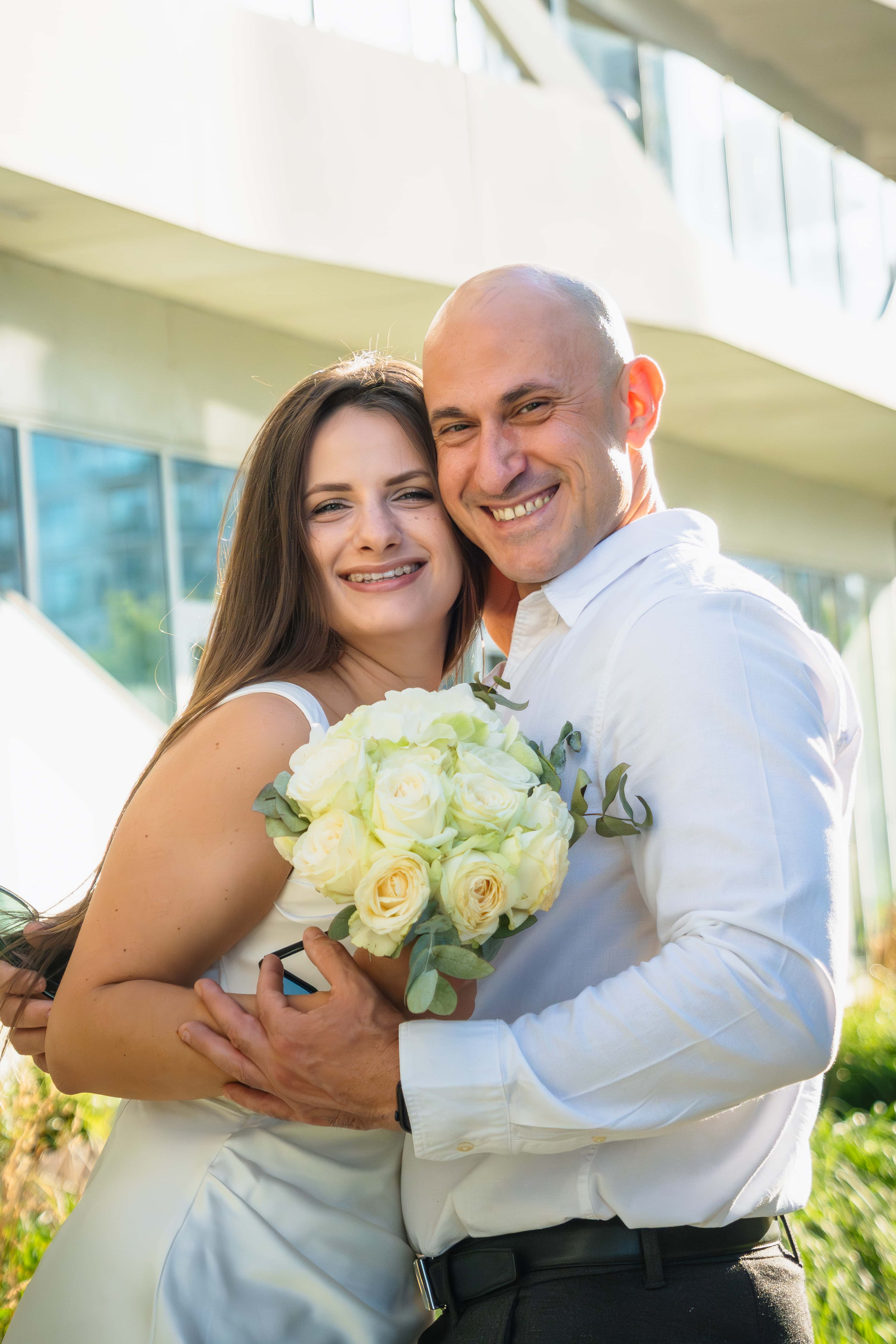 Mariés souriants avec un bouquet de roses blanches, architecture contemporaine, Félix Orain photographe mariage Rennes